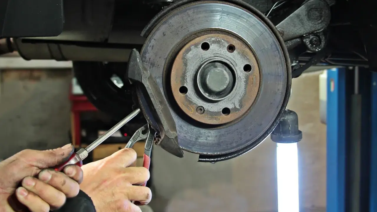 Mechanik wymienia klocki hamulcowe. Widać zużyte tarcze hamulcowe i narzędzia.