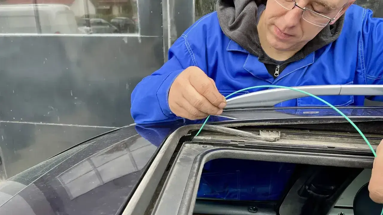 Mechanik w niebieskim kombinezonie pokazuje, jak udrożnić odpływ wody w samochodzie, używając zielonego przewodu do czyszczenia.