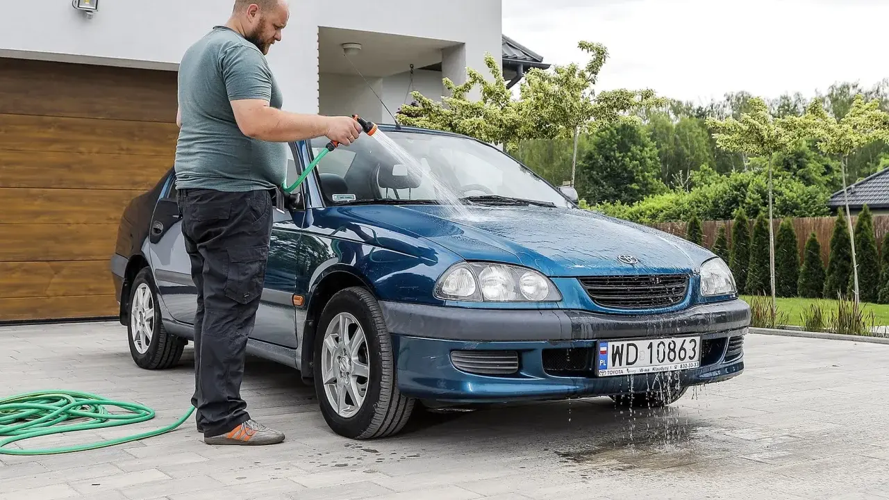 Mężczyzna myje niebieskie auto na posesji, używając węża ogrodowego. Woda spływa po karoserii i tworzy kałuże na podjeździe.
