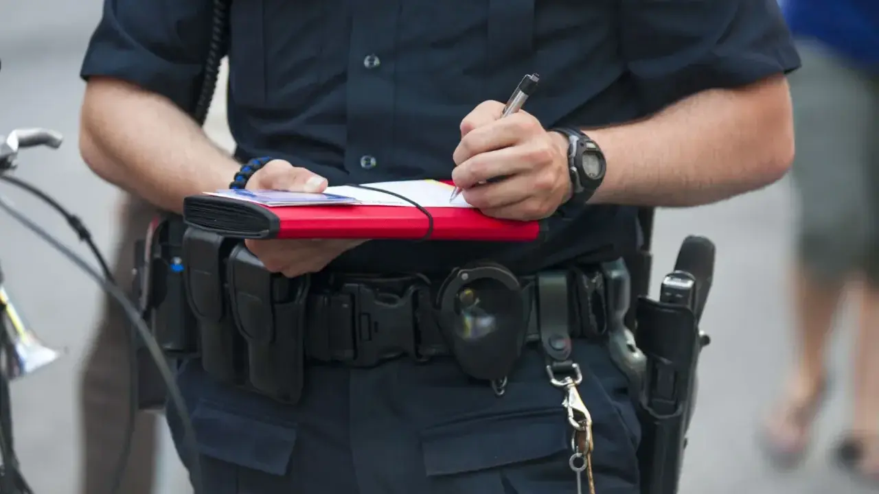 Policjant spisuje mandat, trzymając w ręku notes i długopis. Na pasie ma kaburę z bronią i kajdanki.