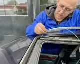 Mechanik w niebieskim kombinezonie pokazuje, jak udrożnić odpływ wody w samochodzie, używając zielonego przewodu do czyszczenia.