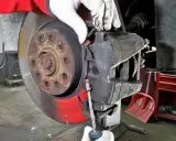 Mechanik w białych rękawiczkach dokonuje wymiany klocków hamulcowych, używając śrubokręta do manipulacji zaciskiem.
