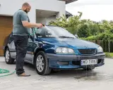 Mężczyzna myje niebieskie auto na posesji, używając węża ogrodowego. Woda spływa po karoserii i tworzy kałuże na podjeździe.
