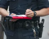 Policjant spisuje mandat, trzymając w ręku notes i długopis. Na pasie ma kaburę z bronią i kajdanki.