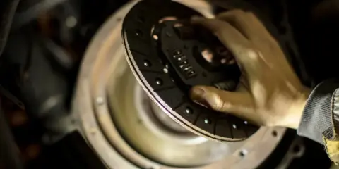 Dłoń trzymająca tarczę sprzęgła nad mechanizmem w samochodzie.