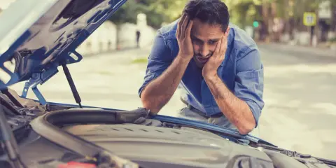Zrozpaczony mężczyzna z rękami na twarzy patrzy na otwartą maskę samochodu. Auto nie odpala, a on nie wie, co robić.