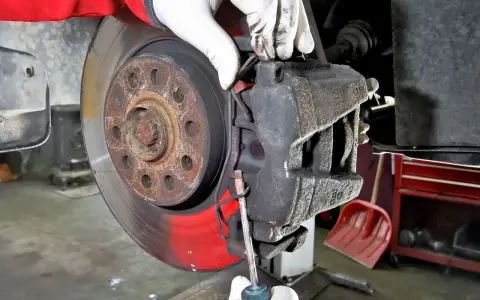 Mechanik w białych rękawiczkach dokonuje wymiany klocków hamulcowych, używając śrubokręta do manipulacji zaciskiem.
