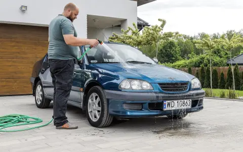 Mężczyzna myje niebieskie auto na posesji, używając węża ogrodowego. Woda spływa po karoserii i tworzy kałuże na podjeździe.