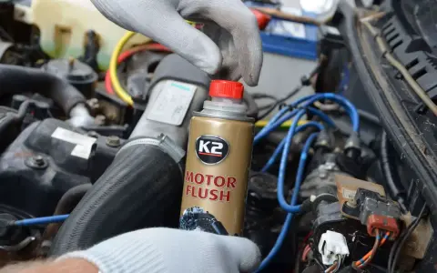Ręka w rękawiczce trzyma puszkę K2 Motor Flush, najlepsza płukanka do silnika benzynowego, gotowa do użycia w samochodzie.