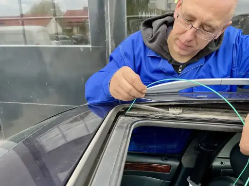 Mechanik w niebieskim kombinezonie pokazuje, jak udrożnić odpływ wody w samochodzie, używając zielonego przewodu do czyszczenia.