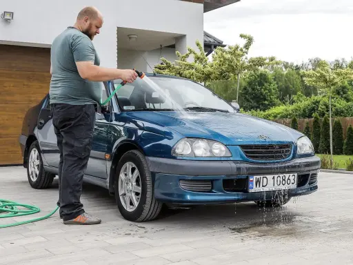 Mężczyzna myje niebieskie auto na posesji, używając węża ogrodowego. Woda spływa po karoserii i tworzy kałuże na podjeździe.