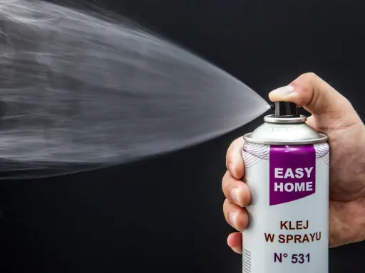Dłoń trzyma puszkę z napisem "EASY HOME KLEJ W SPRAYU", rozpylając białą mgiełkę. Idealny klej do podsufitki bez zdejmowania.