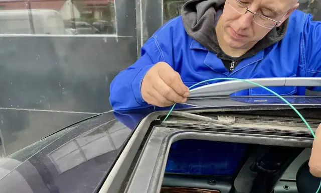 Mechanik w niebieskim kombinezonie pokazuje, jak udrożnić odpływ wody w samochodzie, używając zielonego przewodu do czyszczenia.
