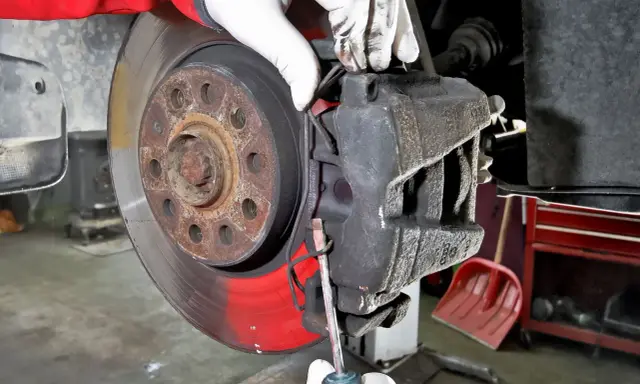Mechanik w białych rękawiczkach dokonuje wymiany klocków hamulcowych, używając śrubokręta do manipulacji zaciskiem.