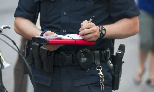 Policjant spisuje mandat, trzymając w ręku notes i długopis. Na pasie ma kaburę z bronią i kajdanki.