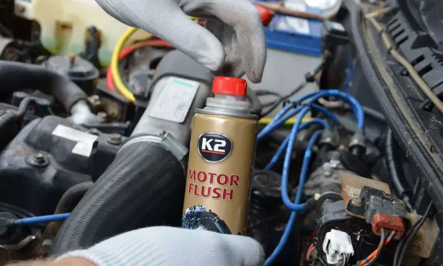 Ręka w rękawiczce trzyma puszkę K2 Motor Flush, najlepsza płukanka do silnika benzynowego, gotowa do użycia w samochodzie.