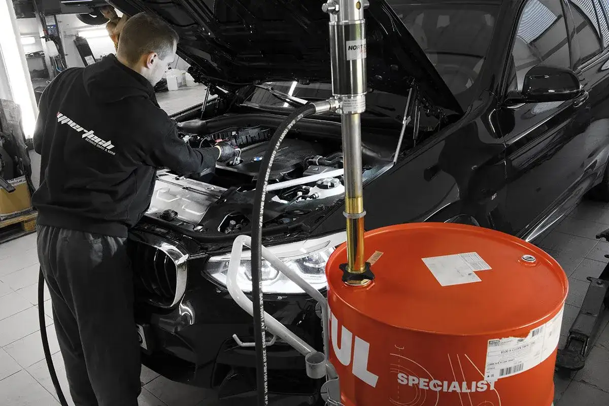 Mechanik wymienia olej w czarnym BMW. Obok stoi duża, pomarańczowa beczka z olejem. Dowiedz się, ile kosztuje wymiana oleju w samochodzie.