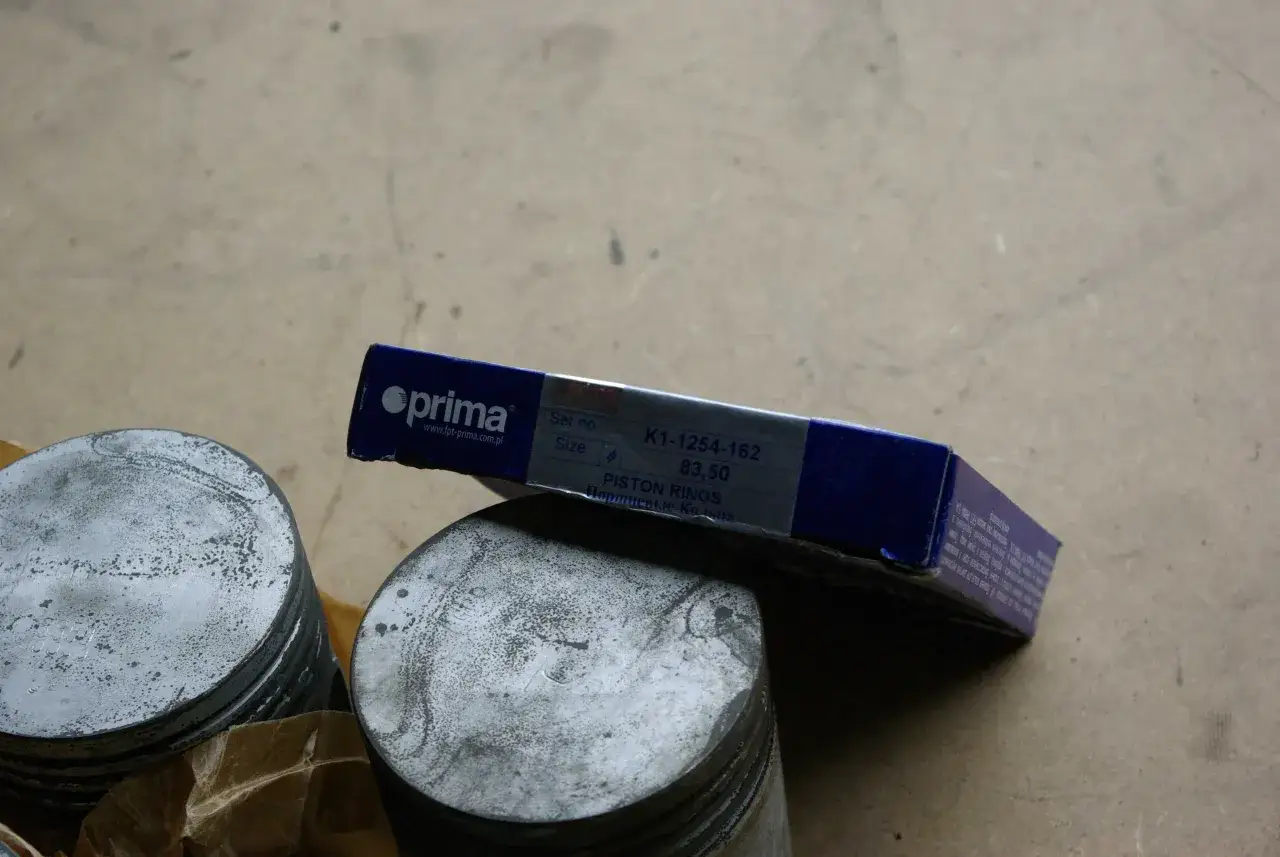Dwa tłoki i pudełko z pierścieniami tłokowymi Prima, z numerem K1-1254-162. To część zamienna, która może pasować do silnika z częściami Kolbenschmidt.