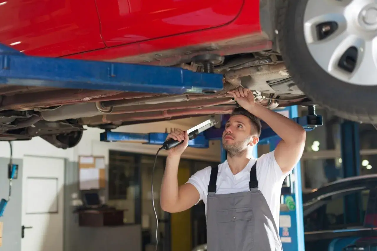 Mechanik z latarką bada podwozie czerwonego auta na podnośniku. Może to być etap przygotowania do kataforezy.