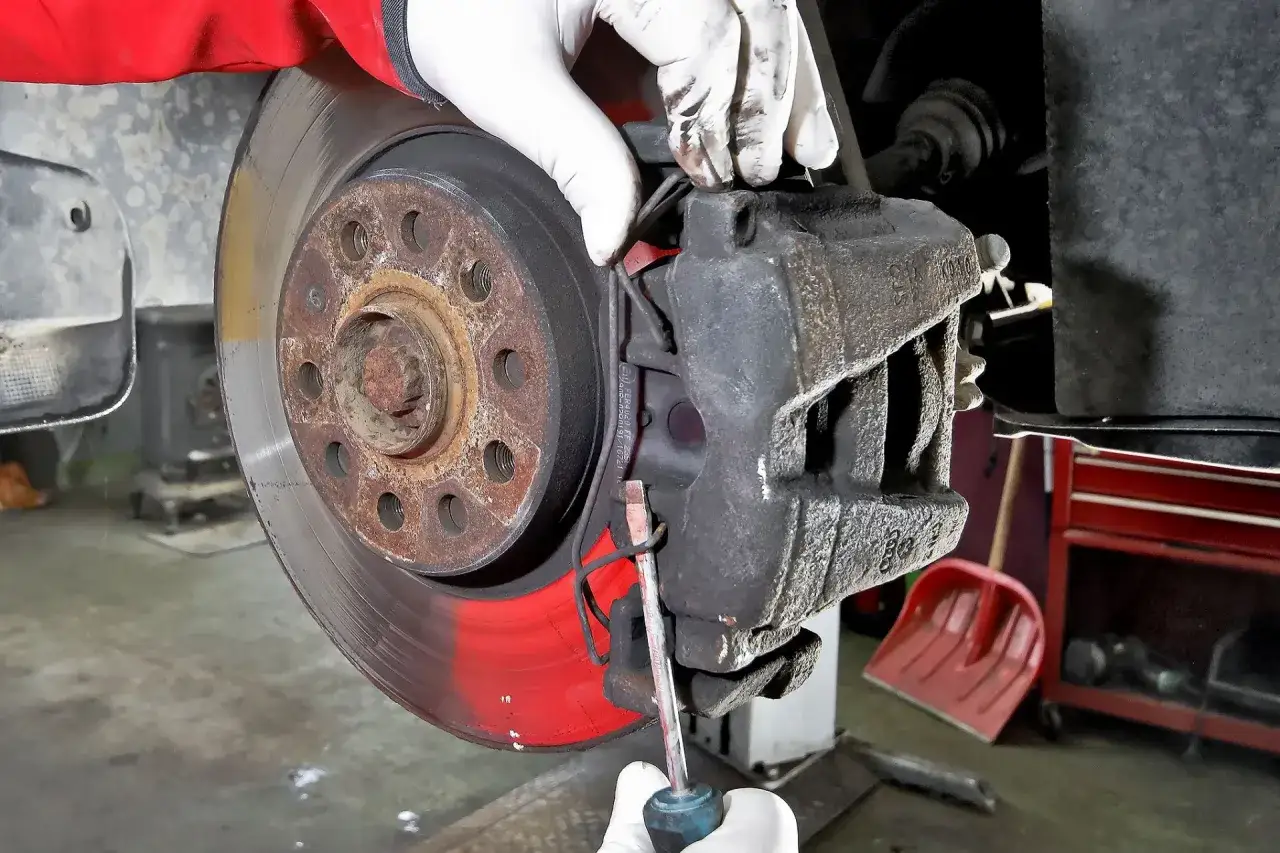 Mechanik w białych rękawiczkach dokonuje wymiany klocków hamulcowych, używając śrubokręta do demontażu.