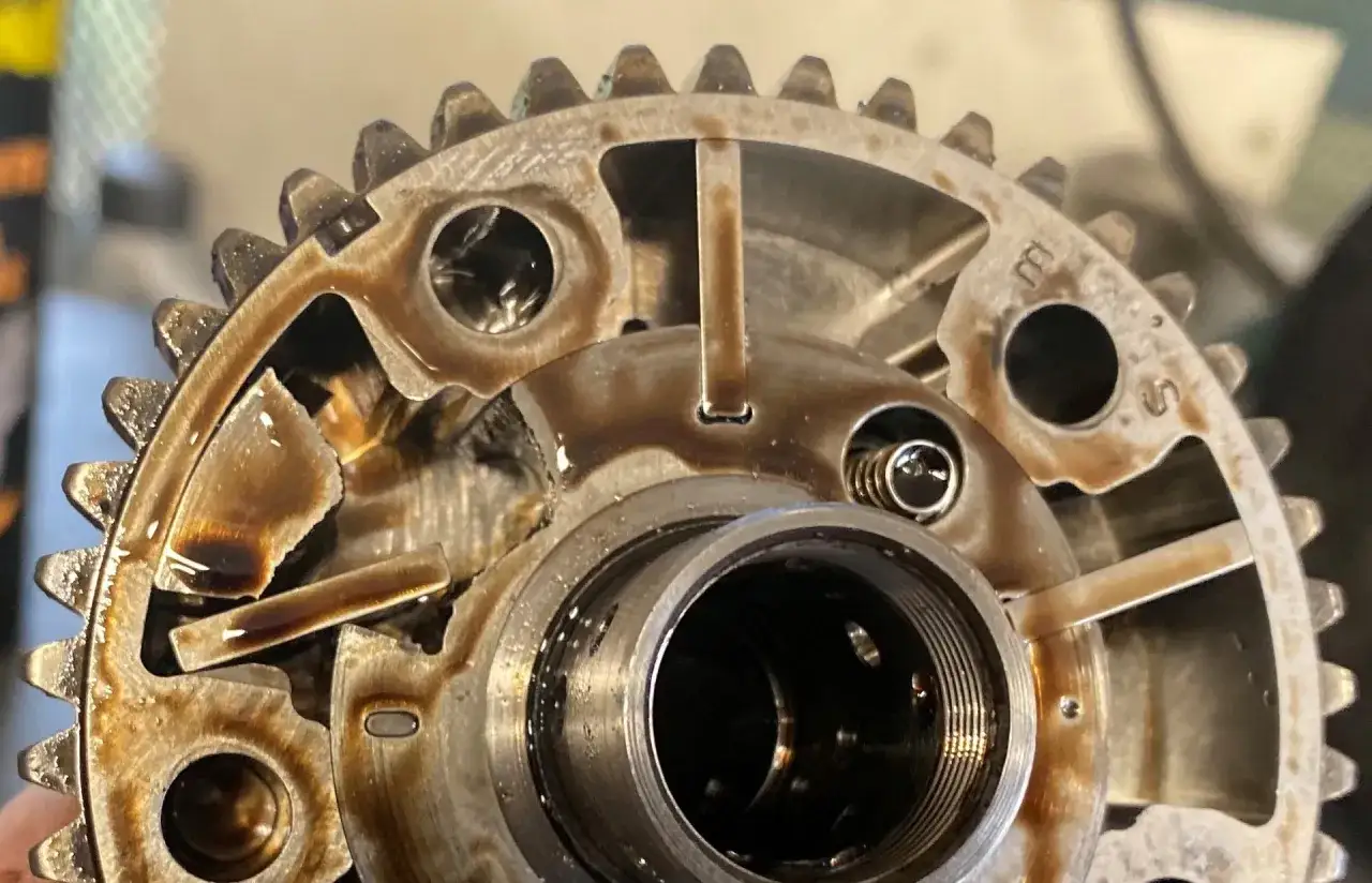 Metalowe koło zmiennych faz rozrządu, pokryte olejem, z widocznymi elementami mechanicznymi i zębatkami.