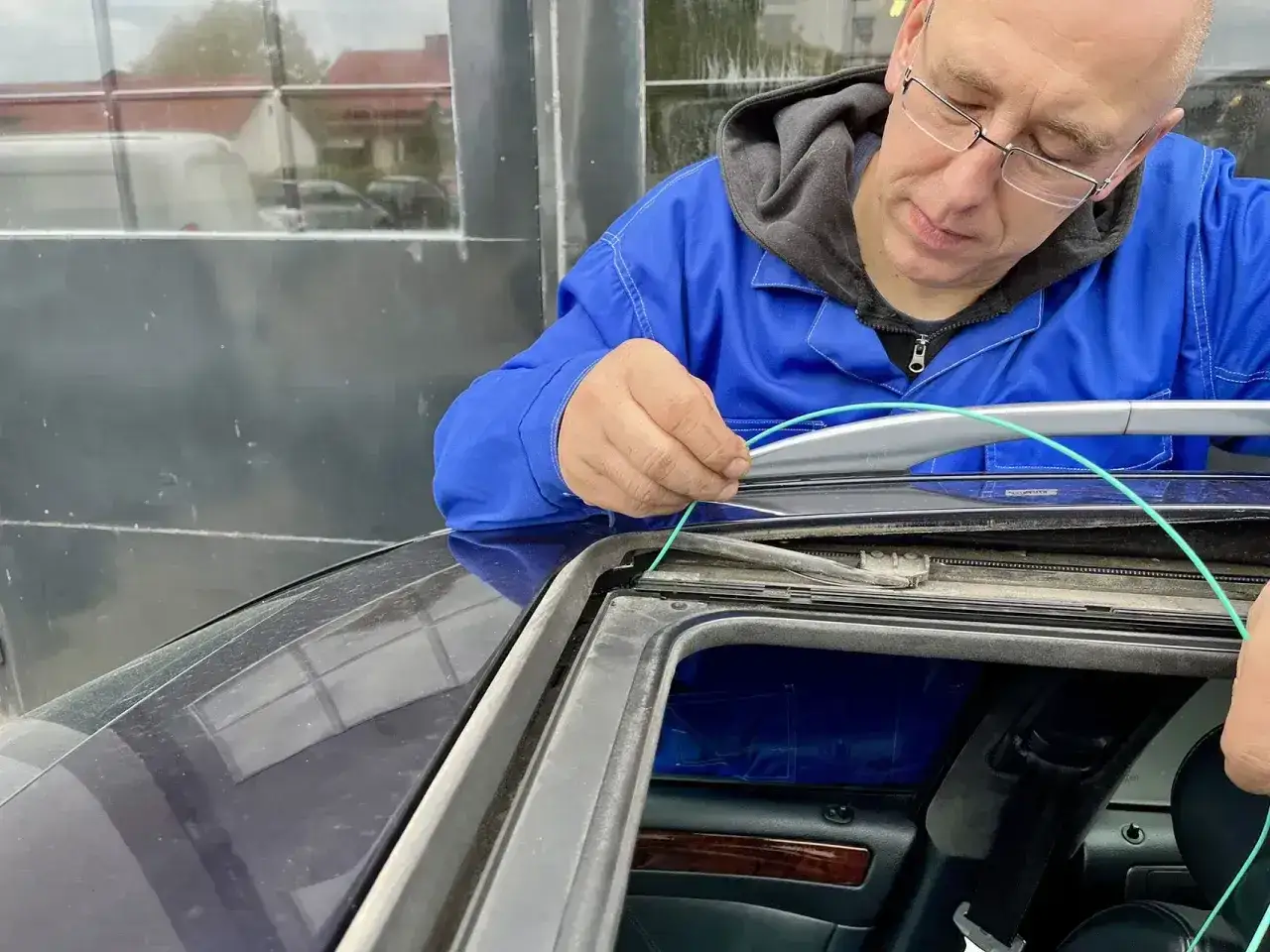 Mechanik w niebieskim kombinezonie pokazuje, jak udrożnić odpływ wody w samochodzie, używając zielonego przewodu do czyszczenia.